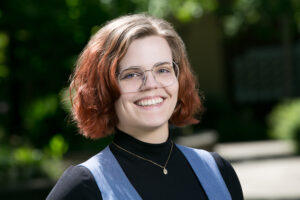 Headshot of Sierra Barnes, a pale-skinned 20-something person with short, reddish hair and wire-framed glasses.