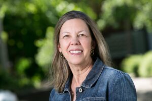 Headshot for Diane Brandsma, Community Support Specialist for Bridge Meadows Beaverton. A pale-skinned woman with long, dark hair peppered with silver. She wears a jean jacket and small loop earrings.