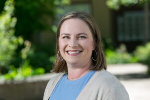 Headshot of Bridge Meadows Research and Evaluation Specialist, Sarah Feldman. She is a pale-skinned woman with shoulder-length brown hair with blonde highlights. She's wearing a sky-blue shirt, ivory cardigan, and large, teardrop shape earrings. 