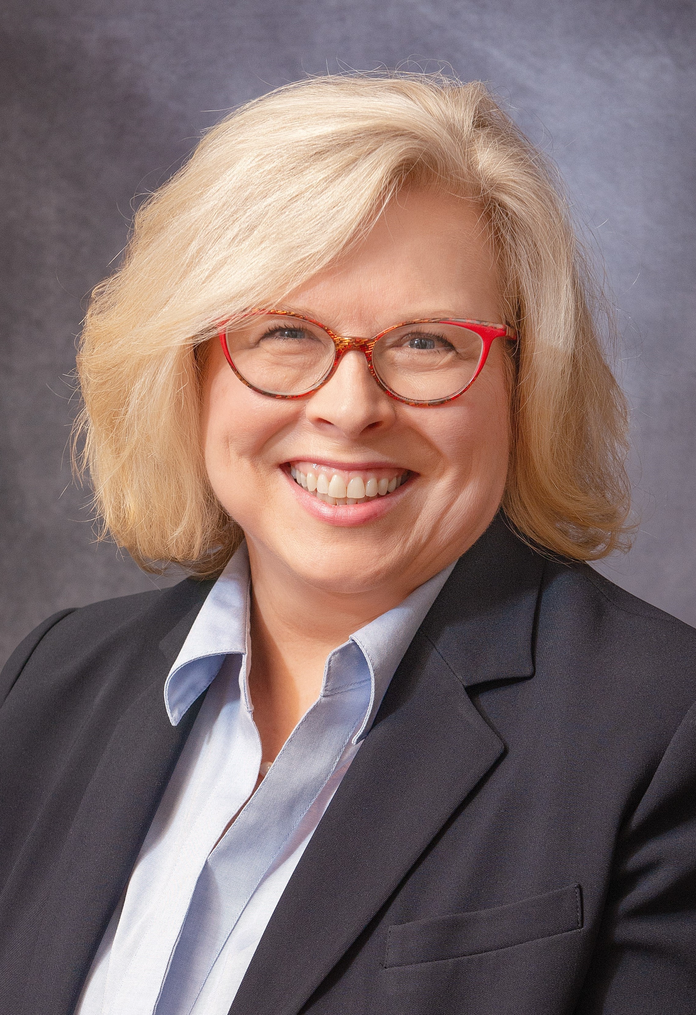 headshot of Executive Director, Dr. Derenda Schubert, a blonde woman wearing red-framed glasses and a dark grey blazer over a pale blue blouse.