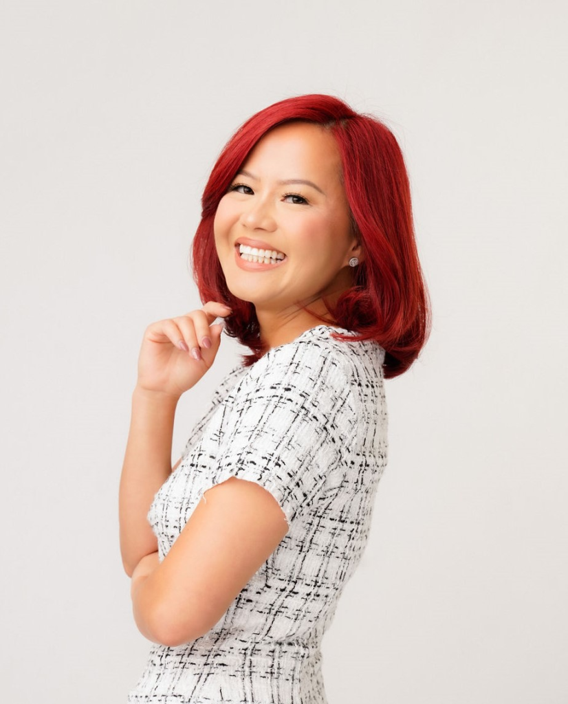 Professional headshot of a young Asian woman with bright red hair and a black and white dress.